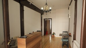 Balcão de madeira na recepção com uma cadeira, um candelabro e um vaso de planta em um corredor com piso de madeira e paredes brancas.