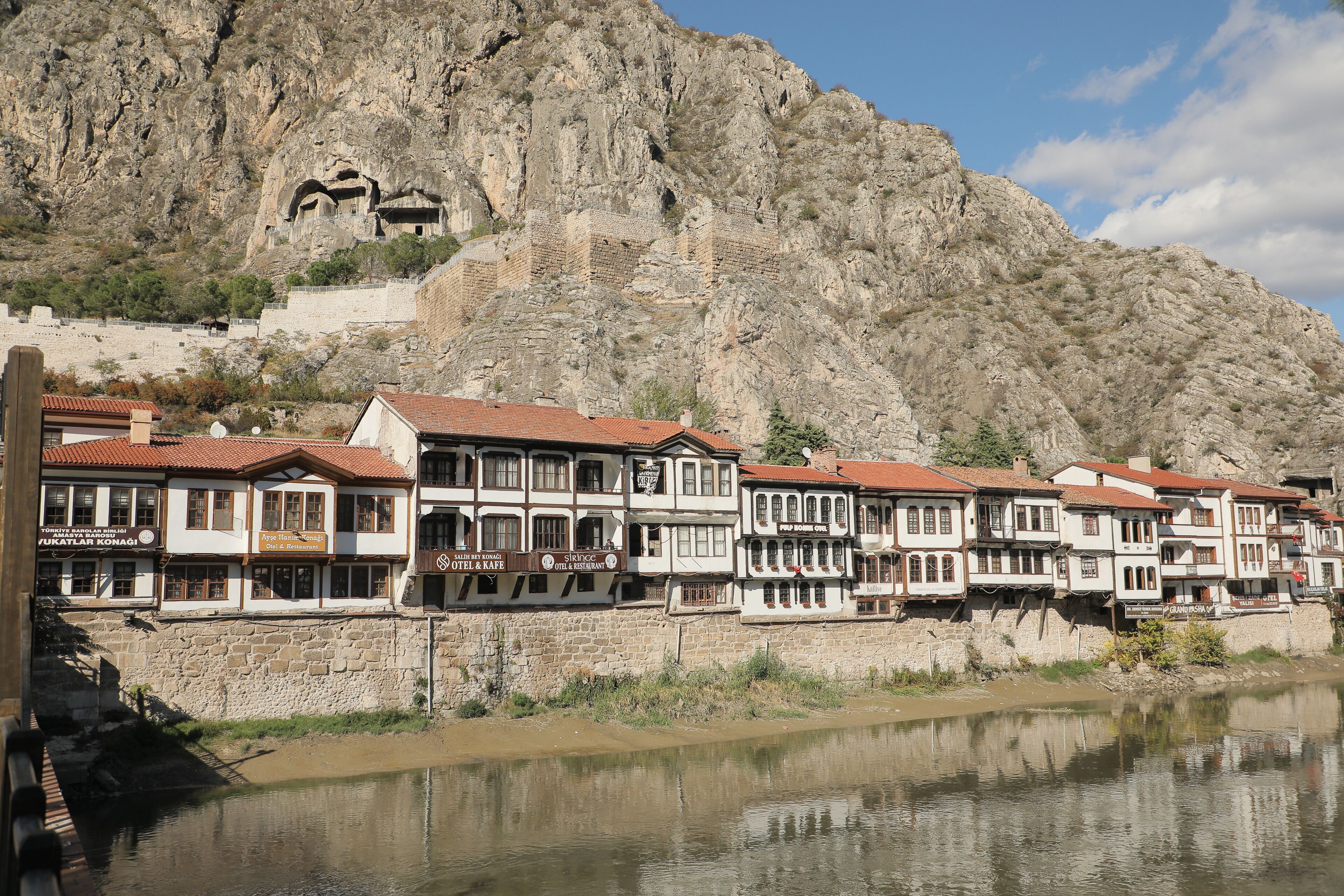 Una hilera de edificios tradicionales con paredes de piedra y techos de tejas rojas, ubicados junto a un río con un telón de fondo rocoso.