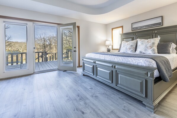 A bedroom with a large bed, a nightstand, a window with a view of trees, and a door leading to an outdoor deck.