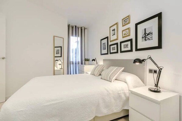 A modern bedroom with a large bed, bedside tables, a desk, and a chair. There are framed pictures on the wall and a window with curtains.