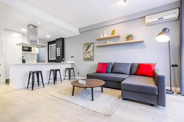 A modern living room with a grey sectional sofa, a round coffee table, and a kitchen area with a white countertop and black stools.