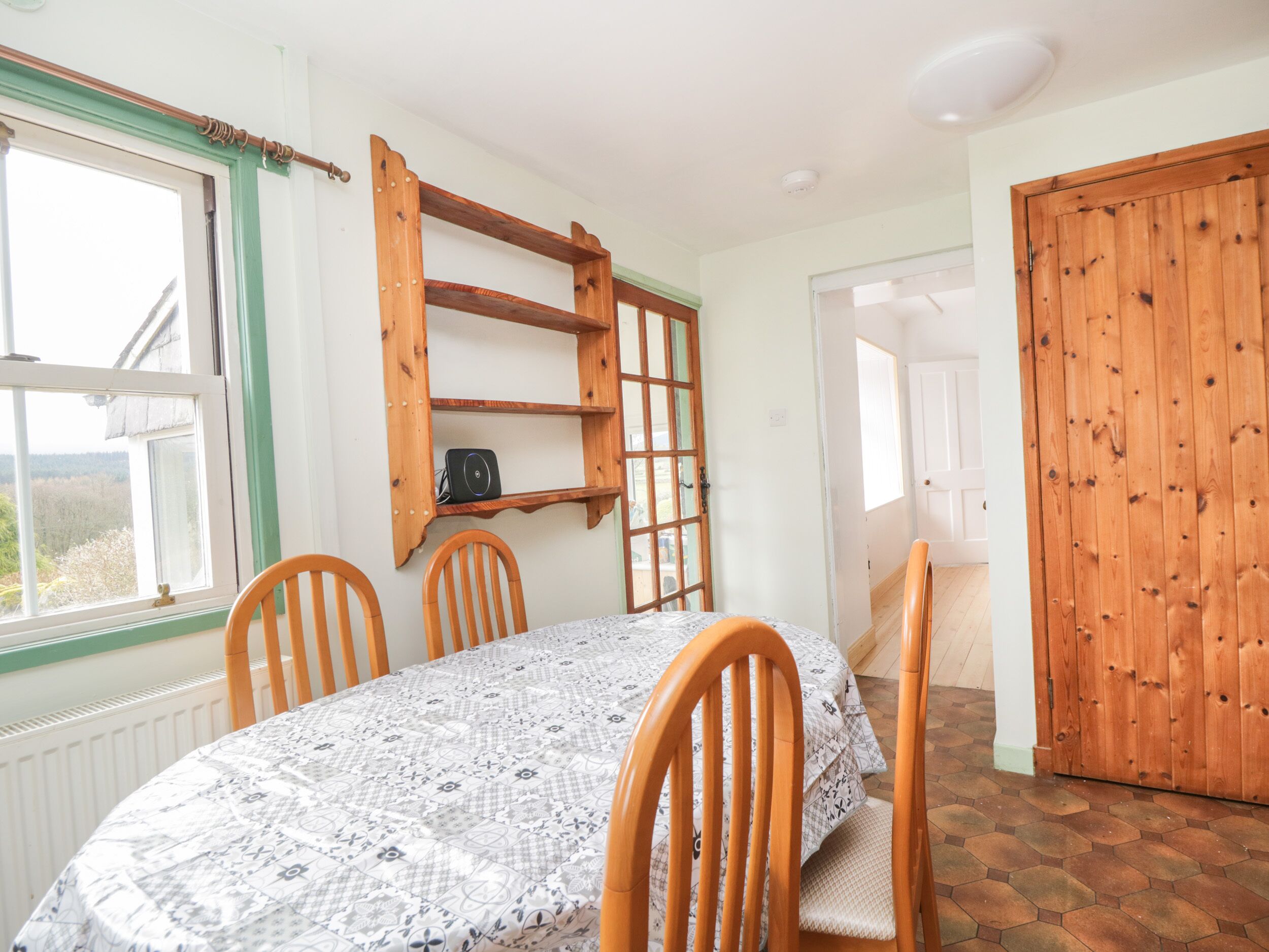 A dining area with a round table covered by a patterned tablecloth, surrounded by wooden chairs. A wooden shelf unit is mounted on the wall above the table.