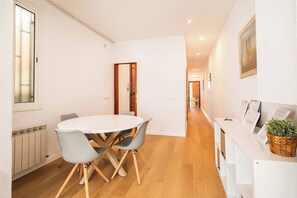 Une chambre d’hôtel moderne avec une table à manger ronde, des chaises grises, un plancher en bois, un radiateur, une porte, un couloir et un comptoir de réception agrémenté d’une plante.
