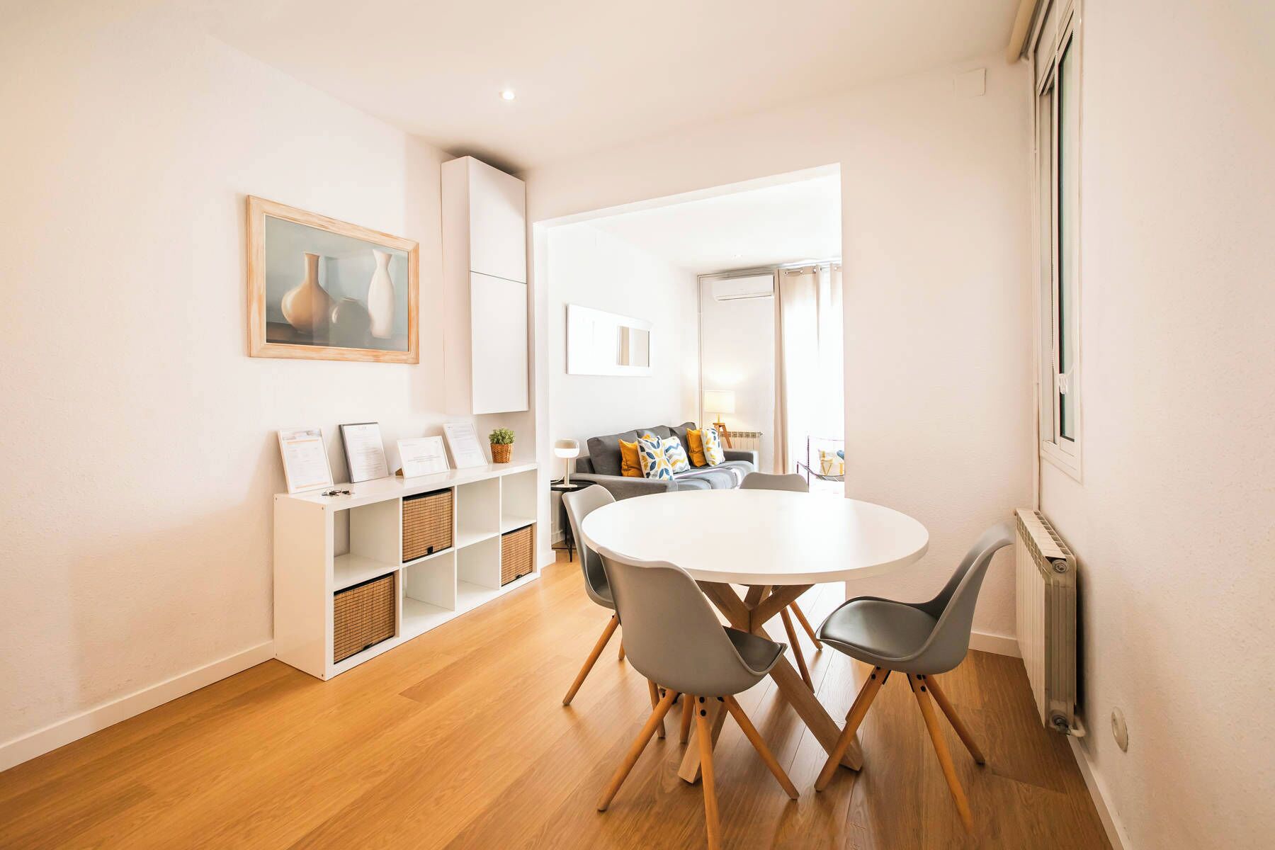 Een moderne woonkamer met een ronde eettafel, grijze stoelen, een grijze bank met gele kussens, een witte kast en een ingelijst schilderij aan de muur.