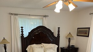 Un dormitorio con una cama grande de madera, dos mesitas de noche con lámparas, un ventilador de techo y una ventana con cortinas.