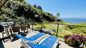 A balcony with two sun loungers, a table, and chairs, overlooking a grassy area and the sea. - Points West Oceanfront Resort 3 bedroom Ocean Point Beach House  (Sooke)