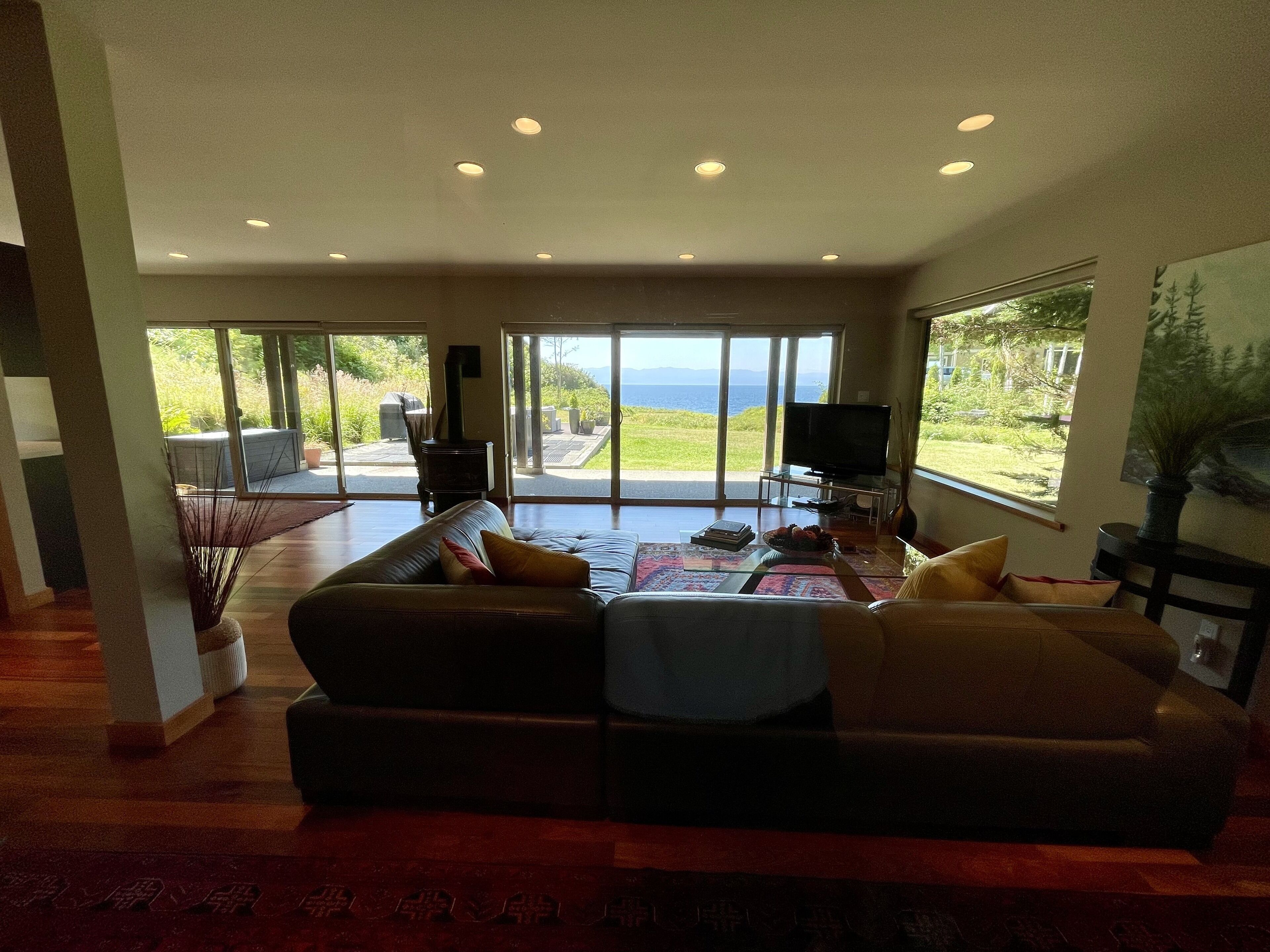 A spacious living room with a large sofa, a coffee table, and a television. There is a fireplace and a view of the outdoors through large windows.