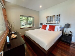 A bedroom with a large bed, a window, a desk with a lamp, and a painting on the wall.