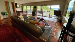 A modern living room with a leather sofa, a glass coffee table, and a large window offering a view of the outdoors. - Points West Oceanfront Resort 3 bedroom Ocean Point Beach House  (Sooke)