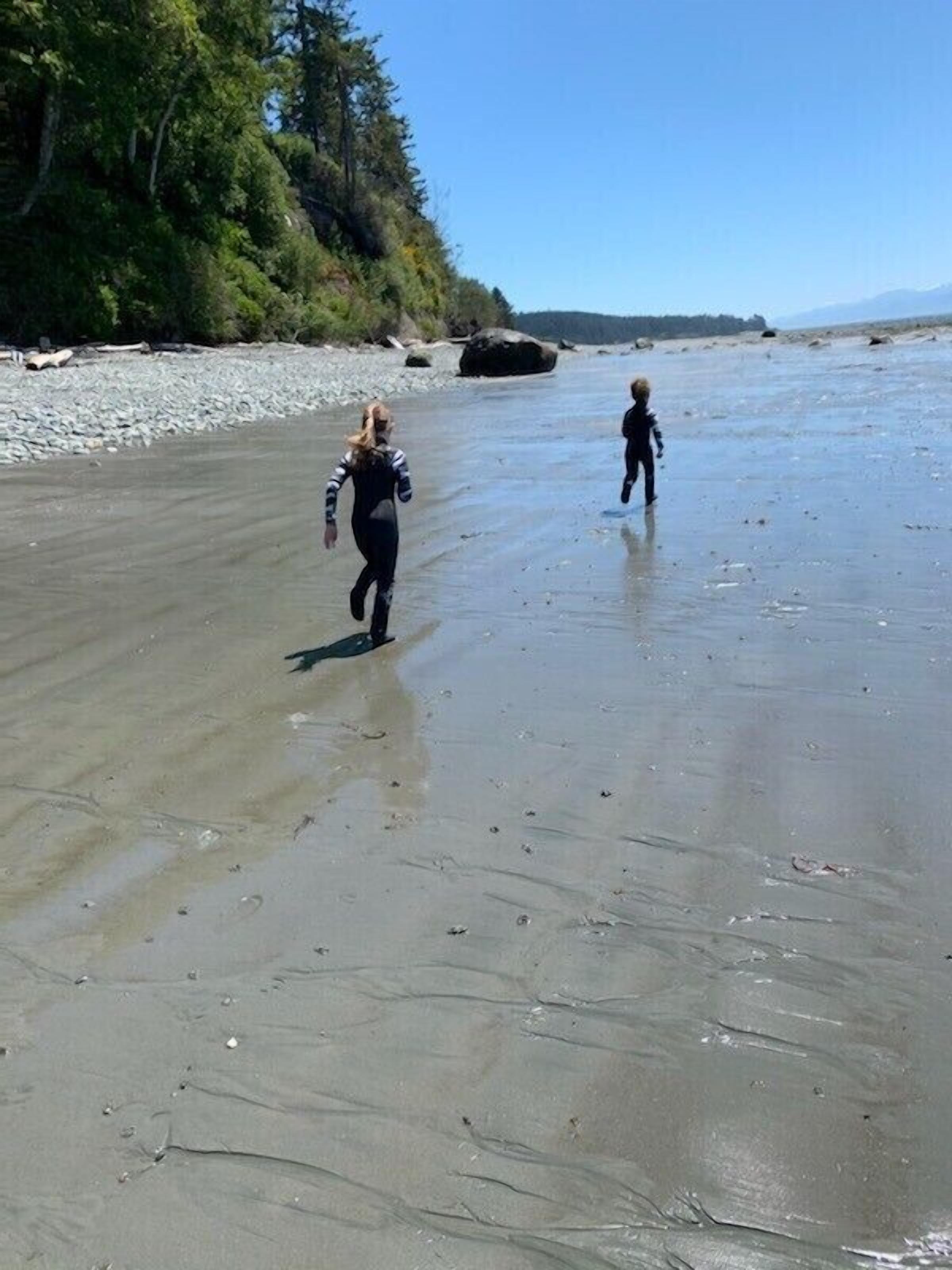 Two people walking on a sandy beach with trees and rocks in the background.