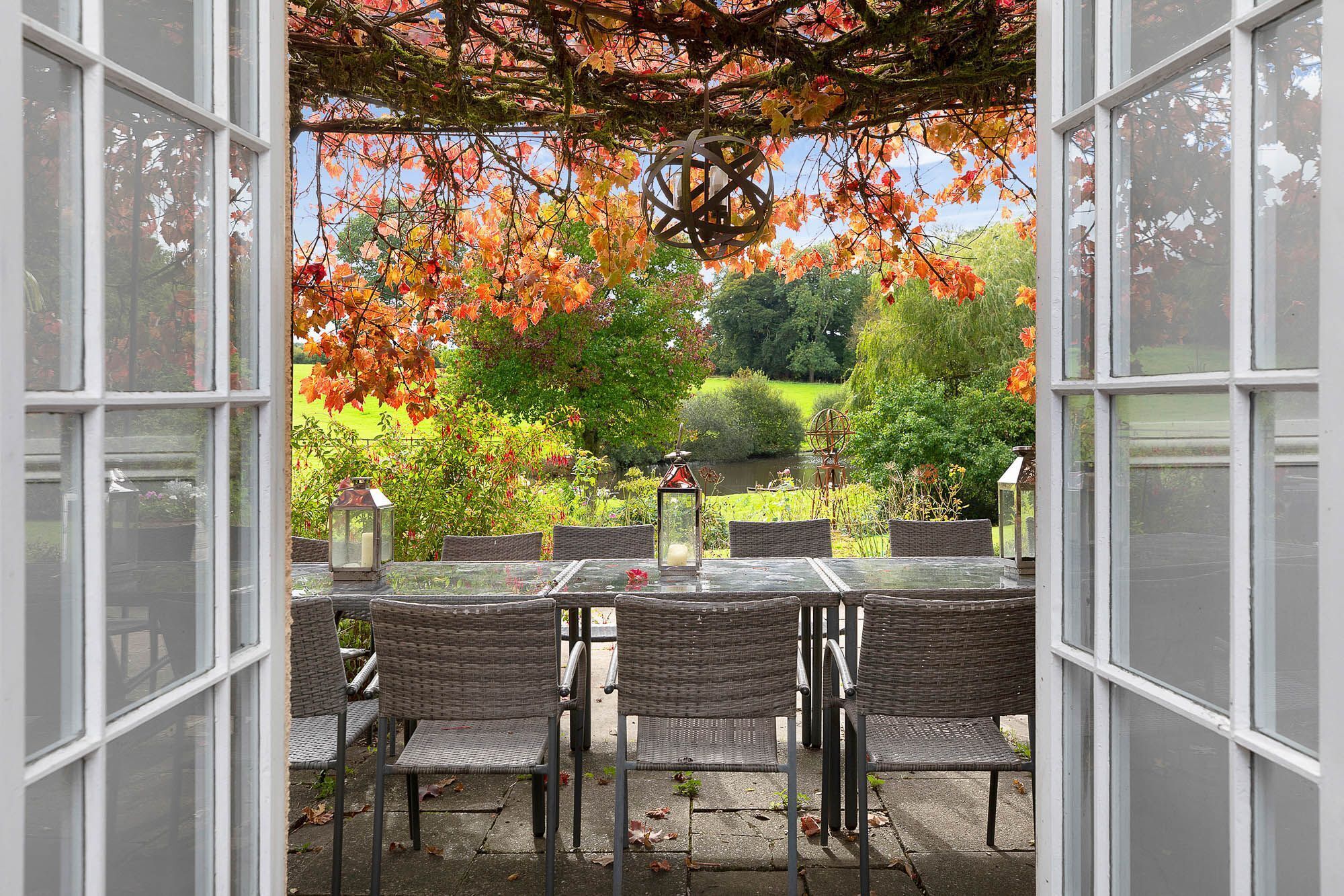 An outdoor patio with a glass table and wicker chairs, surrounded by autumn foliage and garden views.