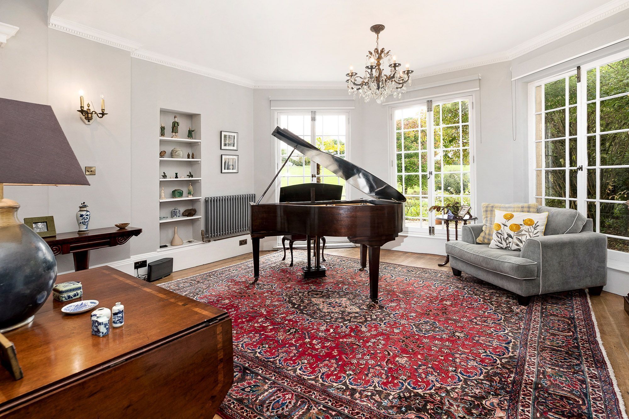 A spacious living room with a grand piano, a grey sofa, and a large window offering a view of greenery.