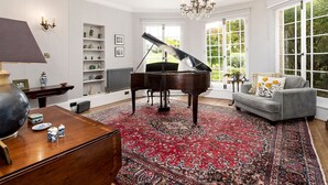 A spacious living room with a grand piano, a grey sofa, and a large window offering a view of greenery. - Colleton Hall - Six Bedroom Villa, Sleeps 12 (Rackenford)