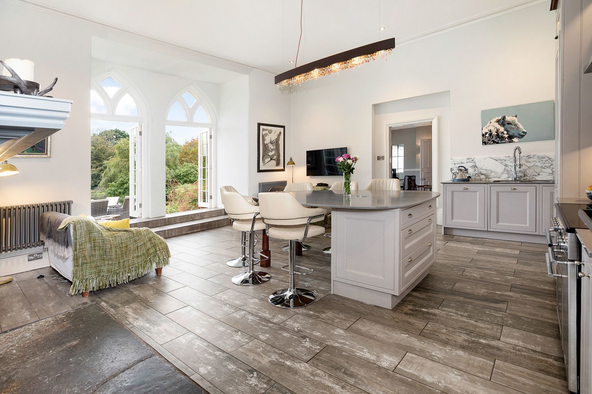 A modern kitchen with a large island, white cabinetry, and a view of the outdoors through large windows.