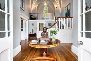 A grand foyer with a wooden staircase, a round table with books and a bust sculpture, and a vase with flowers.