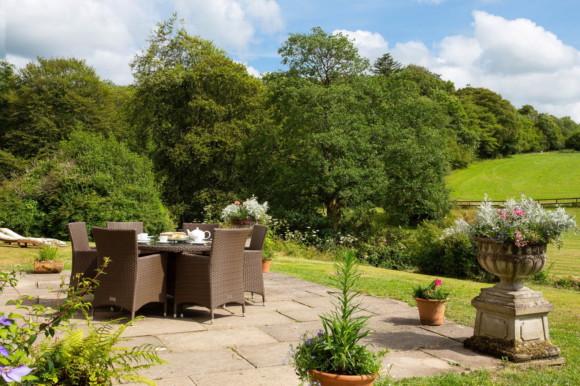 An outdoor dining area with wicker furniture and a table set for tea, surrounded by lush greenery and a stone pathway.