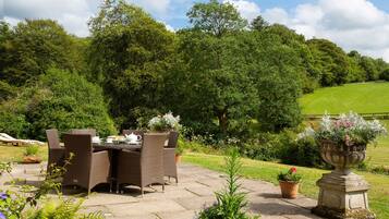 An outdoor dining area with wicker furniture and a table set for tea, surrounded by lush greenery and a stone pathway.
