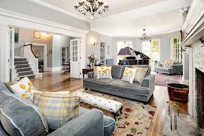 A spacious living room with a grey sofa set, a grand piano, and a chandelier. - Colleton Hall - Six Bedroom Villa, Sleeps 12 (Rackenford)