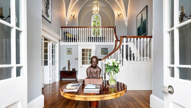 A grand staircase with white balustrades leading to a second floor, a round wooden table with books and a vase of flowers, and a statue on the table.