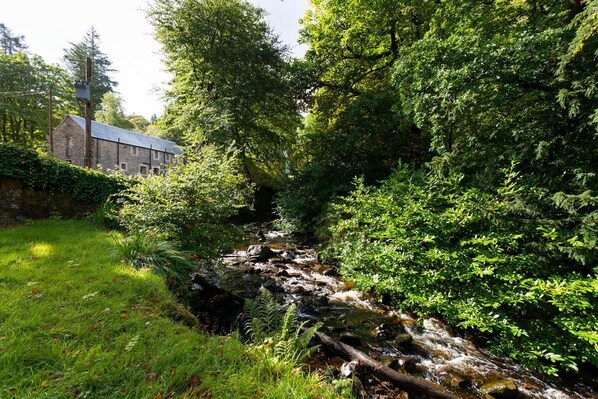 A stone building with a sloped roof, surrounded by lush greenery and a flowing stream.