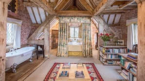 Une chambre mansardée au style rustique, avec des poutres apparentes, une grande fenêtre ornée de rideaux à motifs floraux, une baignoire ancienne et une bibliothèque remplie de livres.