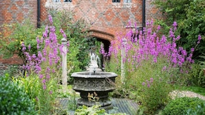Un jardin agrémenté d’une fontaine en pierre, entouré d’une végétation luxuriante et de fleurs violettes, devant un bâtiment en briques doté de fenêtres en arc de cercle.