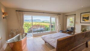 A bedroom with a large bed, a wooden bench, a bathtub, and a balcony with a view of the countryside. - Derwent Garden Estate (Bridekirk)