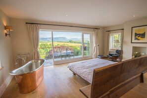 A bedroom with a large bed, a wooden bench, a bathtub, and a balcony with a view of the countryside. - Derwent Garden Estate (Bridekirk)