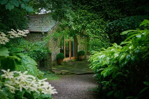A stone building with a wooden door, surrounded by lush greenery and a gravel path leading to it. - Derwent Garden Estate (Bridekirk)