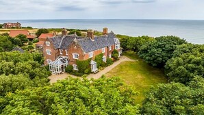 A large brick house with multiple chimneys, surrounded by lush greenery and a view of the sea. - Meadow Manor - Seven Bedroom Villa, Sleeps 14 (Mundesley)