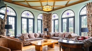 A spacious living room with a large sofa set, a coffee table, and a round table. The room has large arched windows with curtains, wooden flooring, and a high ceiling with a chandelier. - Culver Down Manor - Seven Bedroom Villa, Sleeps 16 (Shanklin)