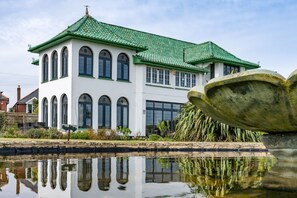 A traditional building with a green roof and arched windows, reflecting in a calm water body in front.