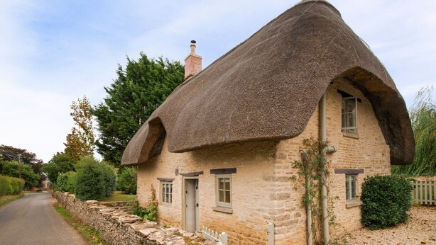 A traditional stone cottage with a thatched roof, a stone wall, and a narrow road leading to it.