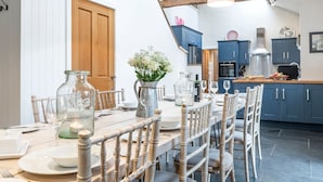 Comedor con una mesa de madera dispuesta para cuatro personas, rodeada de sillas. Hay botellas de vidrio y un florero con flores sobre la mesa. La habitación tiene techo de madera y se observa una cocina al fondo con armarios azules.