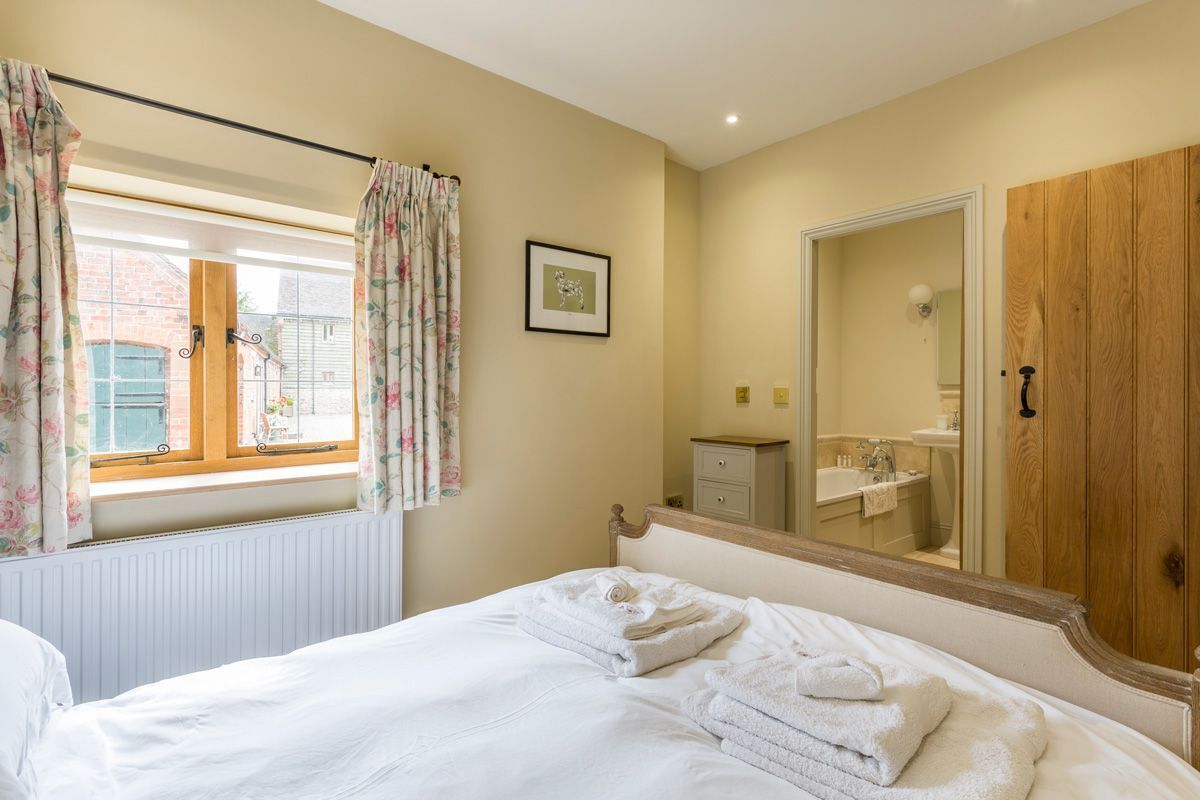 Une chambre à coucher avec un lit, une fenêtre avec des rideaux, une table de chevet, un miroir et une armoire en bois.