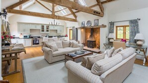 A spacious living room with a high ceiling and exposed wooden beams, a fireplace, and a view of the kitchen in the background. - Chevaliers Moat House (Upton Cressett)