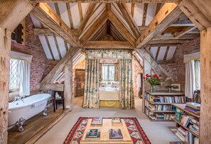 Une chambre mansardée au style rustique, avec des poutres apparentes, une grande fenêtre ornée de rideaux à motifs floraux, une baignoire ancienne et une bibliothèque remplie de livres.
