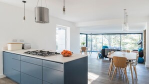 A modern kitchen with a central island, a dining area with a table and chairs, and large windows offering a view of the outdoors.