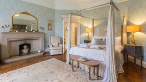 Une chambre à coucher avec un lit à baldaquin, un coin salon avec un fauteuil, une cheminée et une table de chevet avec une lampe.