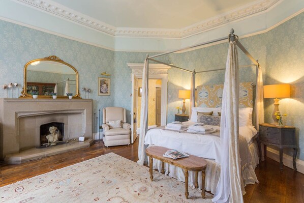 Une chambre à coucher avec un lit à baldaquin, un coin salon avec un fauteuil, une cheminée et une table de chevet avec une lampe.