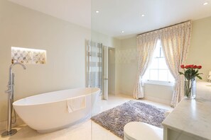 Une salle de bain moderne avec une baignoire sur pieds, une douche en verre, un lavabo en marbre et une fenêtre agrémentée de rideaux à motifs floraux.