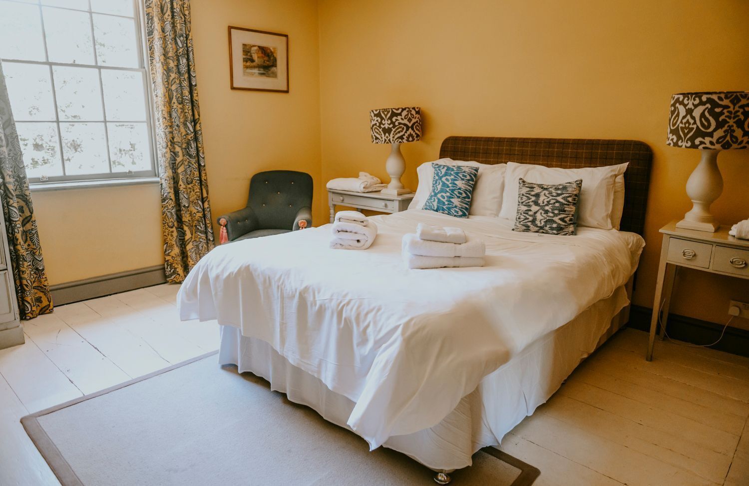 A neatly made bed with white linens and a few decorative pillows, a chair, a bedside table with a lamp, and a window with curtains.