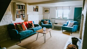 Uma sala de estar aconchegante com um sofá azul, uma poltrona combinando e uma mesa de centro. Uma estante está encostada à parede, e uma janela oferece iluminação natural.