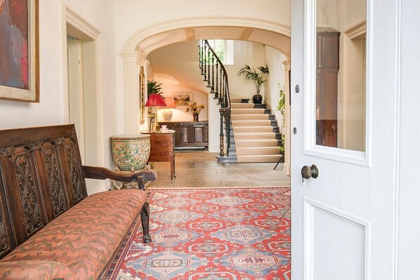 A hallway with a patterned rug, a wooden bench, a decorative vase, and a staircase leading to a second floor.