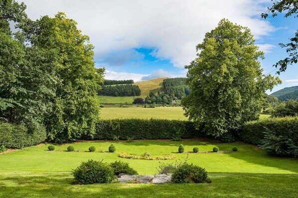 Een verzorgde tuin met verschillende bomen en struiken, een strak kort gazon en een helder uitzicht over het omliggende landschap.