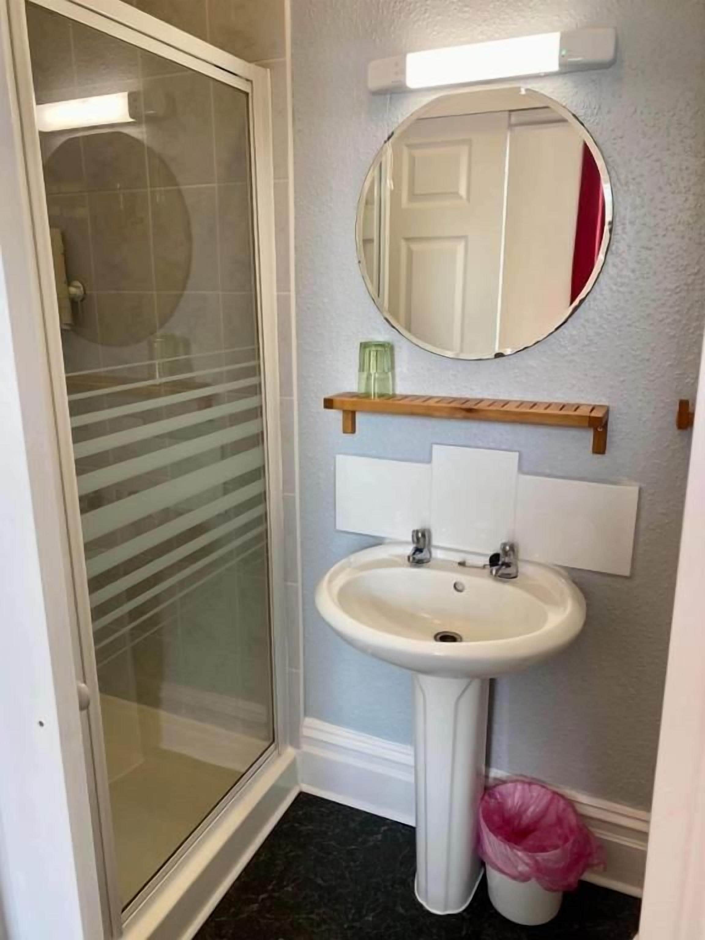 Een badkamer met een douchecabine van glas, een wastafel op voet, een spiegel en een afvalbak.