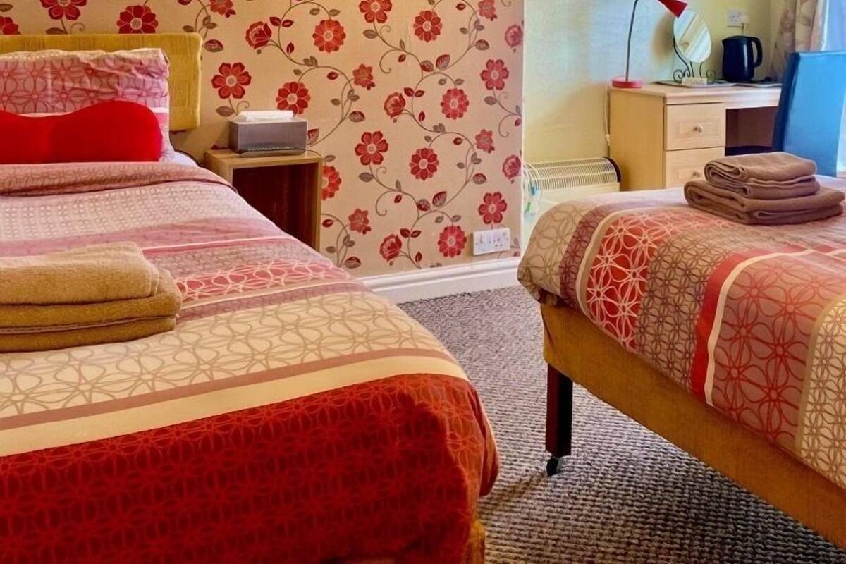 Un dormitorio con papel tapiz floral, una cama con ropa de cama roja y beige, una mesita de noche con una lámpara y un escritorio con una silla.