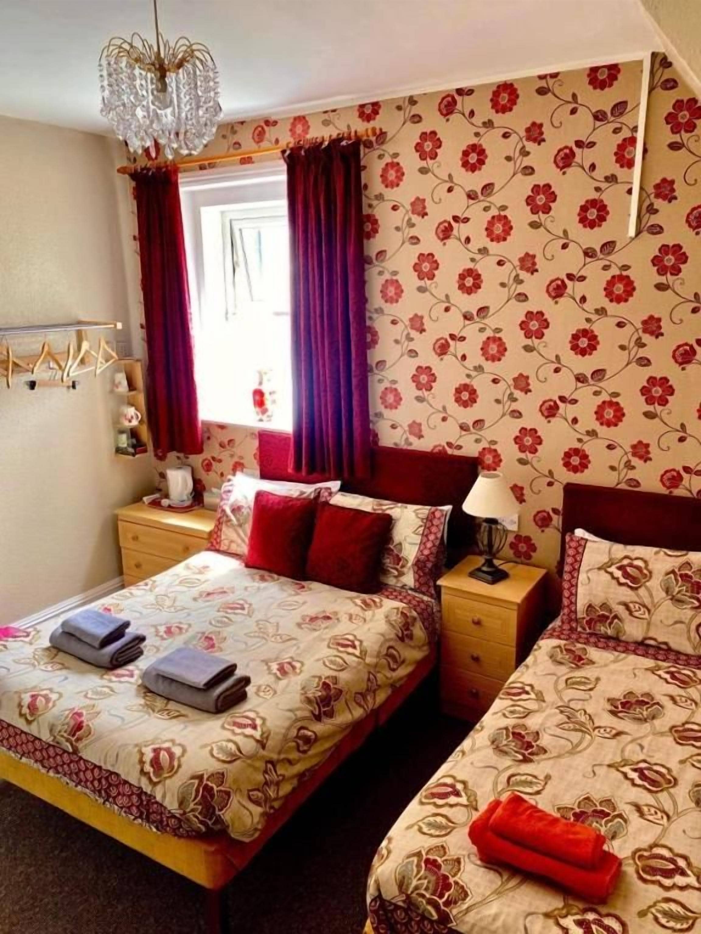 A bedroom with two beds, a chandelier, a window with curtains, and a floral wallpaper design.