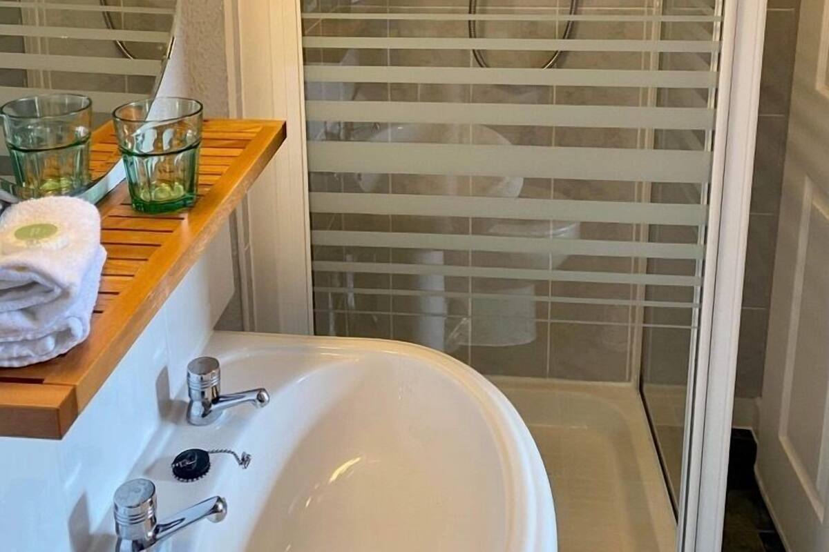 Un baño con ducha de vidrio, lavamanos blanco y estante de madera con vasos y toalla.