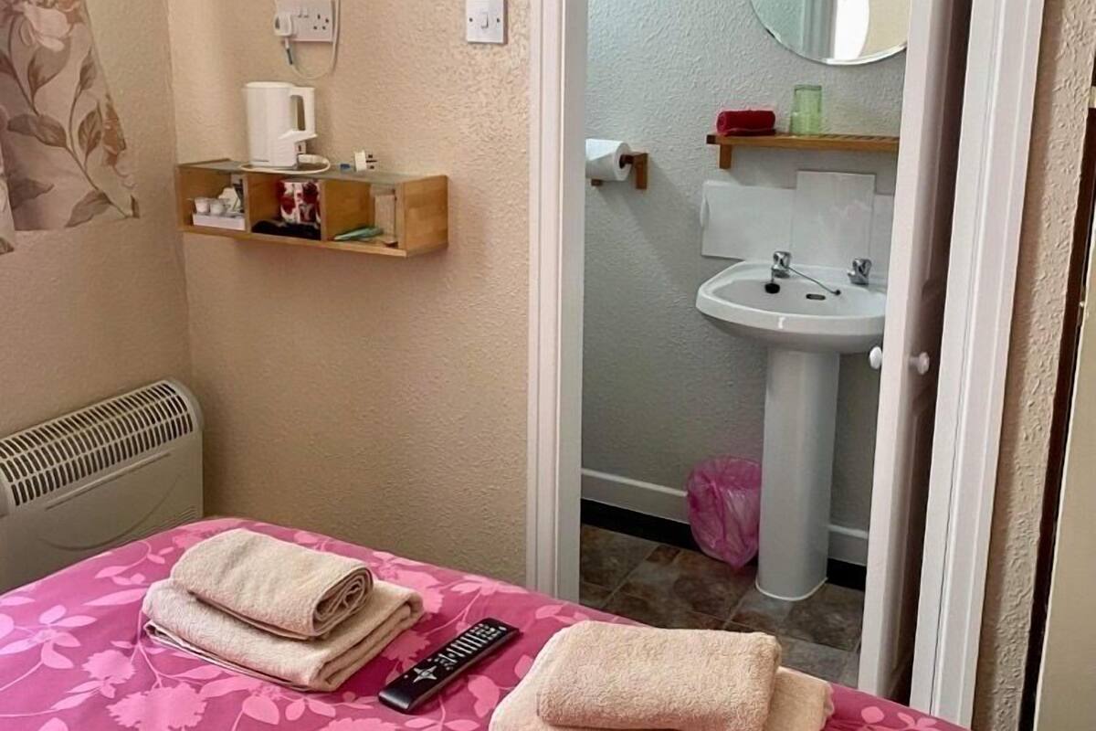 Un dormitorio con una cama, un televisor montado en la pared, una mesita de noche con cafetera y un baño visible a través de una puerta abierta.